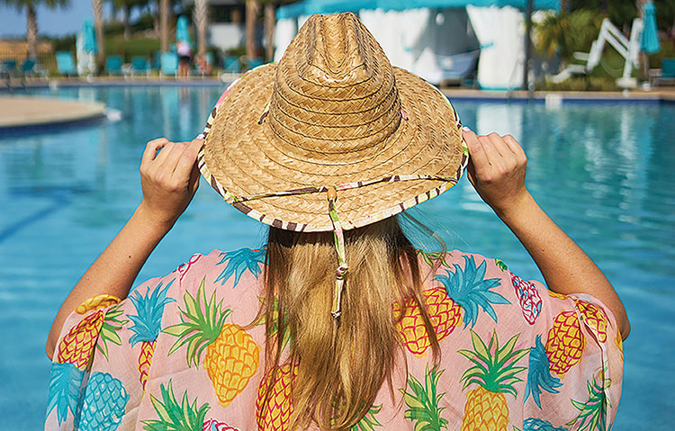 Woman wearing a straw hat and pineapple and pink printed dress overlooking a rich blue pool