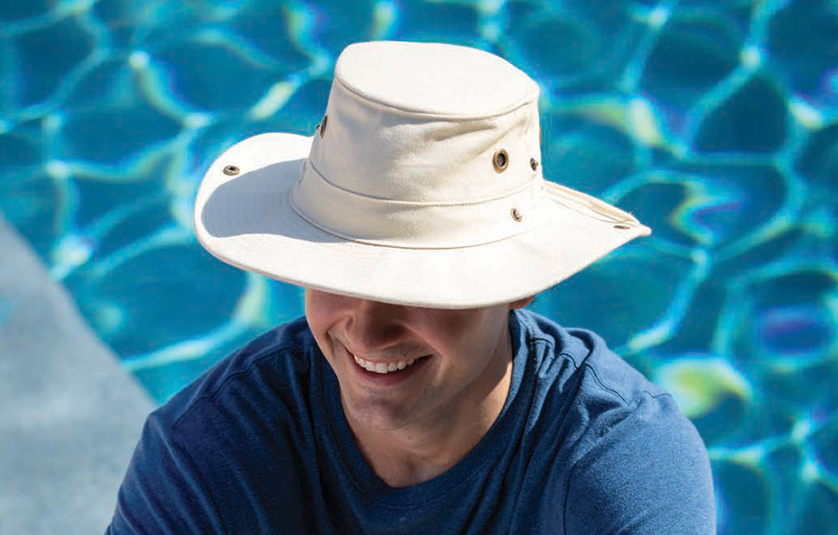 Man wearing a white hat and blue longsleeve shirt with brass airholes embedded into the hat sitting near a glassy-blue pool