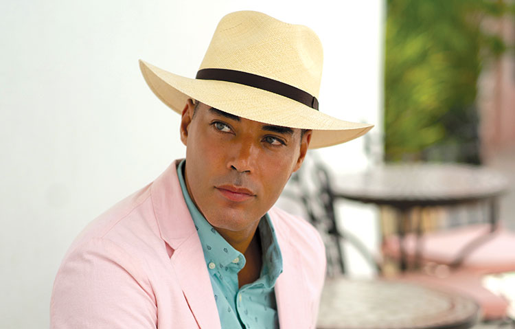 Man wearing a light tan hat with dark brim and a pink suit and light blue shirt while sitting near some tables