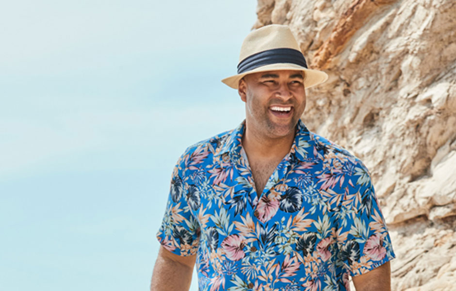 Smiling man wearing a tan hat with black ribbon in a hawaiian shirt against a yellowed  rocky mountain