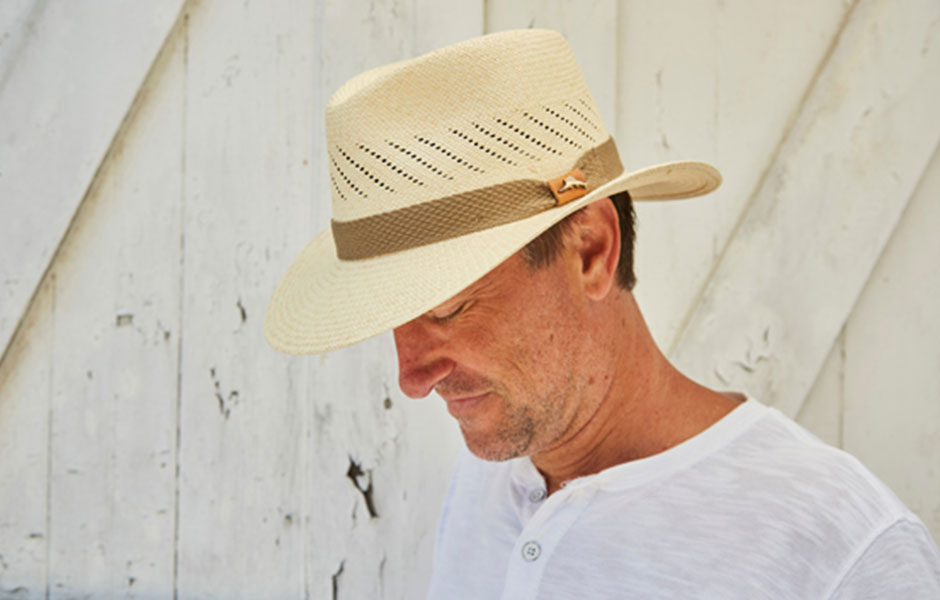 Man wearing a tan hat with a brown ribbon and white shirt looking down towards the ground leaning against a white wall