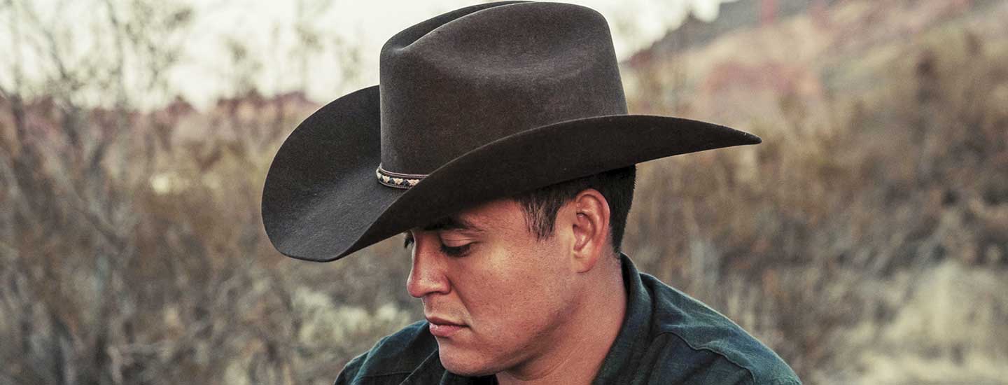 Man wearing a brown cowboy hat and green sweatshirt looking down in a dry field