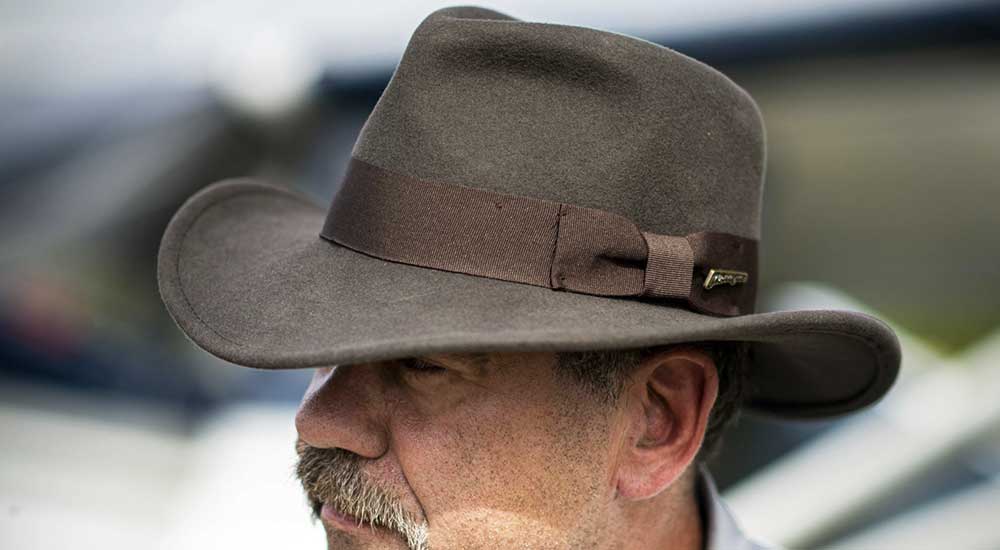 Older man with a grey mustache wearing a dark grey and brown Indiana Jones hat