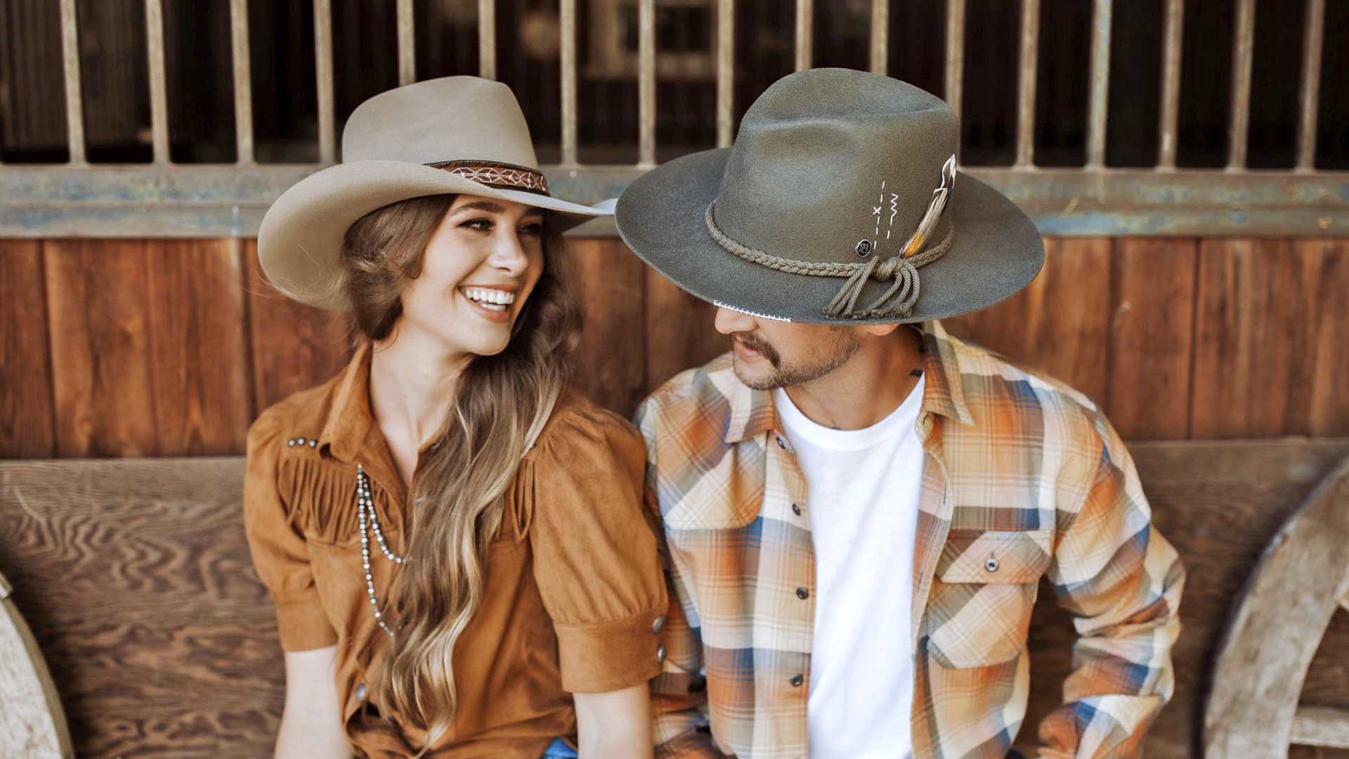 Woman and man smiling at each other wearing cowboy hat and attire leaning against a horse stable